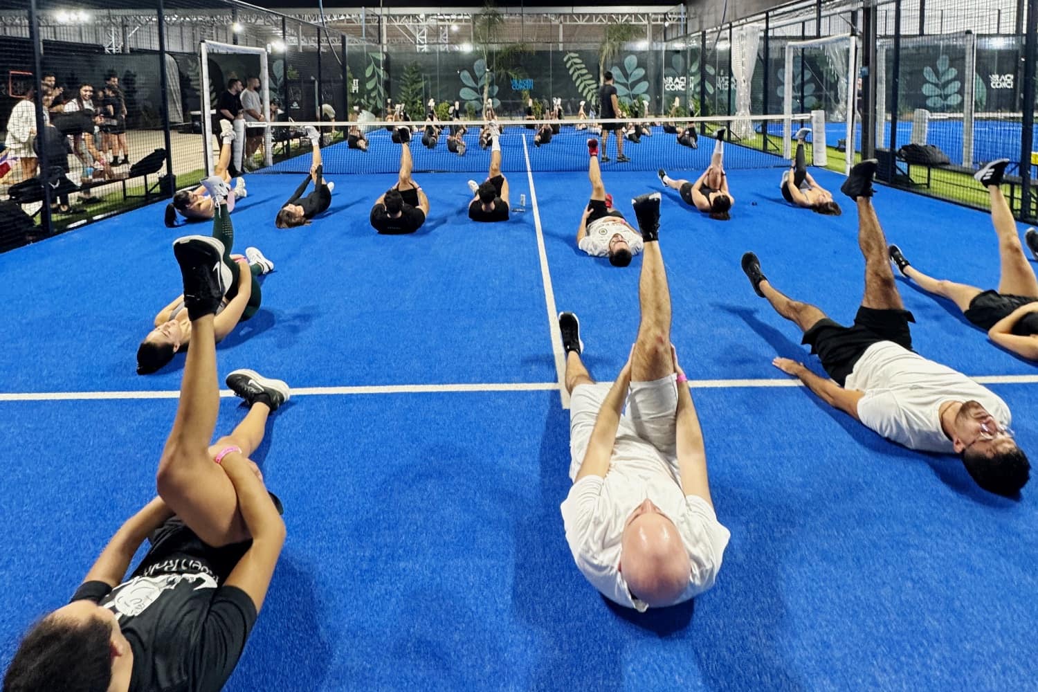 The whole group lying across a floodlit blue padel court at night, legs raised through an exercise