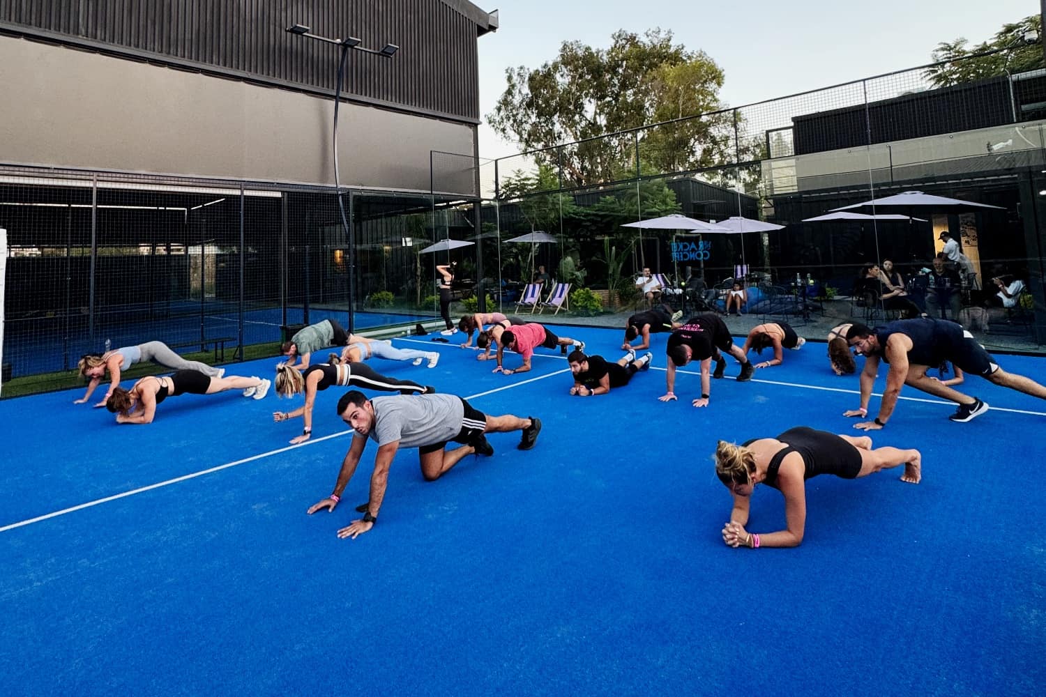 A large group stretching together on a blue padel court under an evening sky