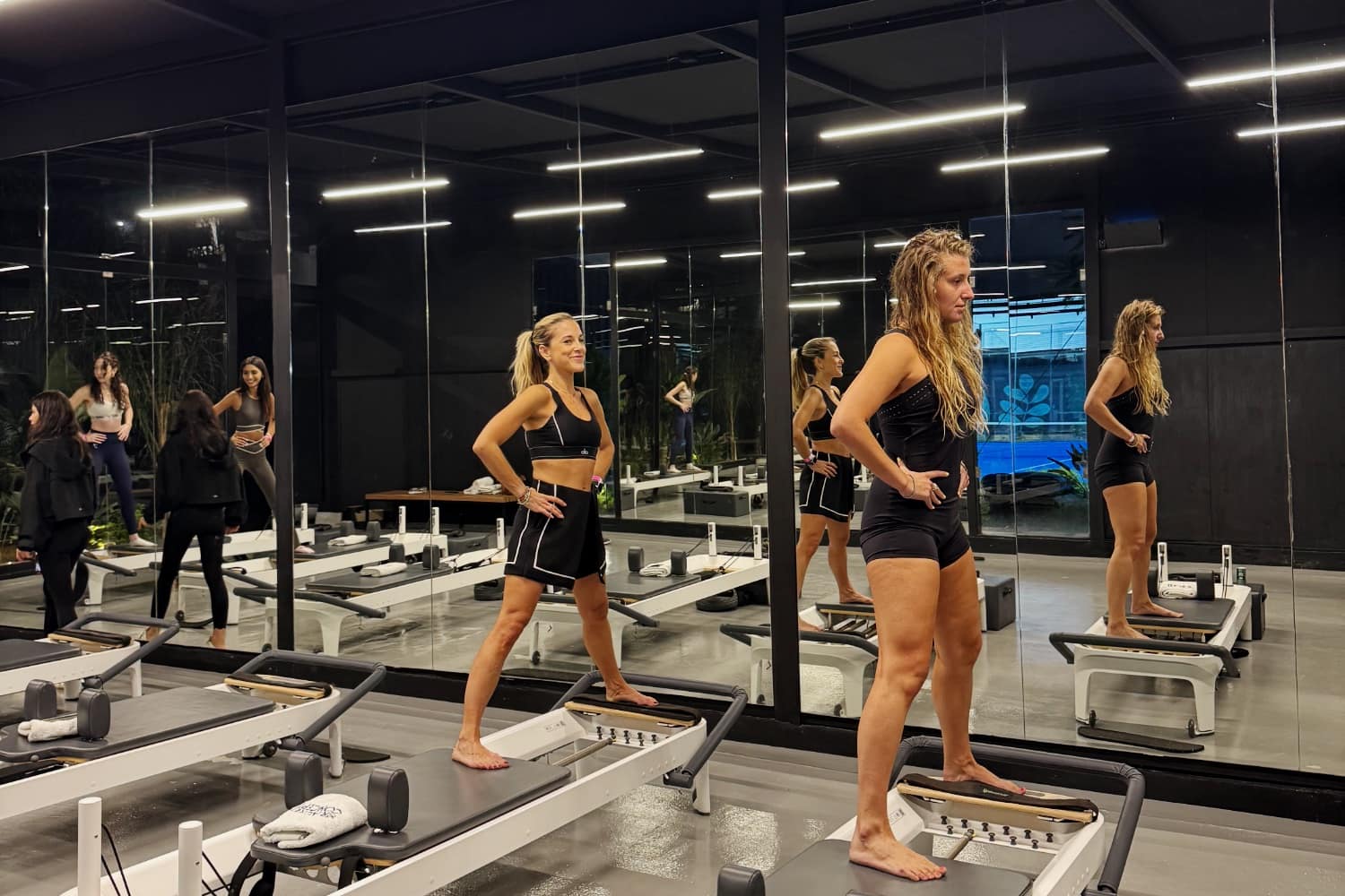 Two members standing on reformer machines in a mirrored studio, the class reflected behind them
