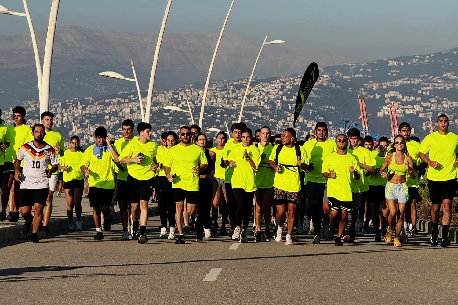 A wide field of Run Club Beirut runners in club yellow filling the corniche, the mountains behind them
