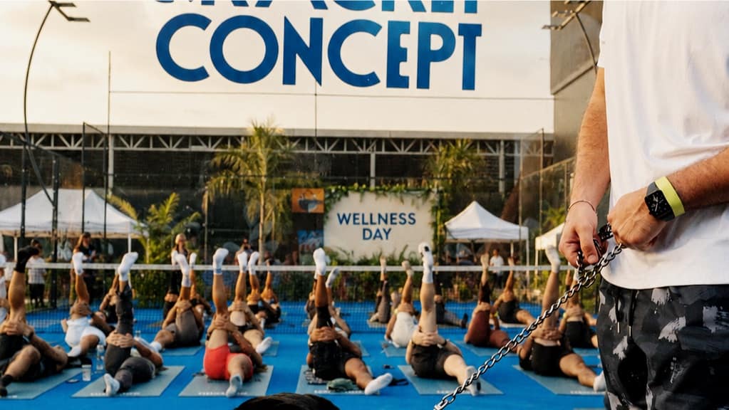 A Wellness Day class stretched out across the courts at The Racket Concept, the venue sign above them and a member walking a dog past the mats